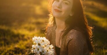 A woman smiles while holding white flowers in a sunlit outdoor setting.