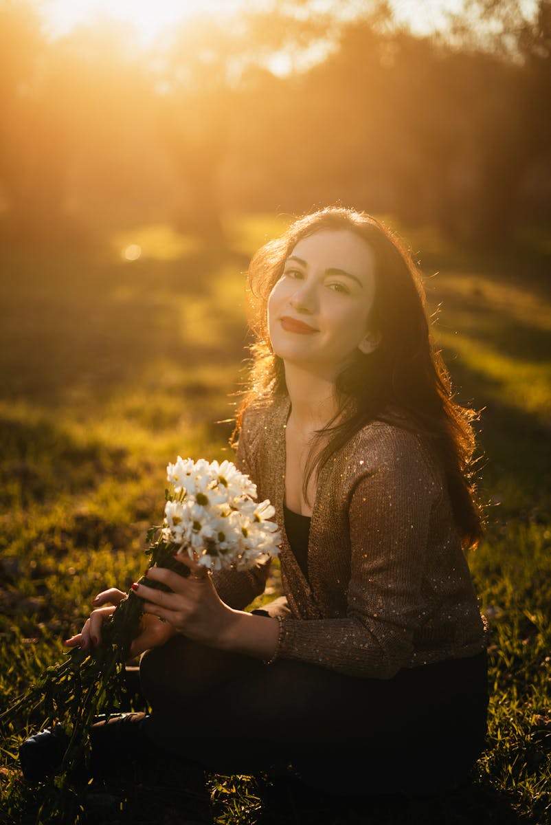 A woman smiles while holding white flowers in a sunlit outdoor setting.