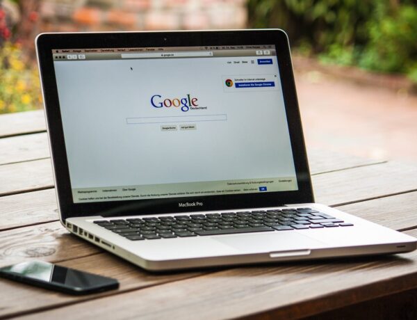 A MacBook Pro displaying Google Search on a wooden table outdoors, next to a smartphone.